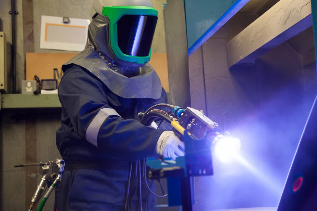 Welder in protective gear and helmet using a welding torch, with a bright arc in an industrial workshop.