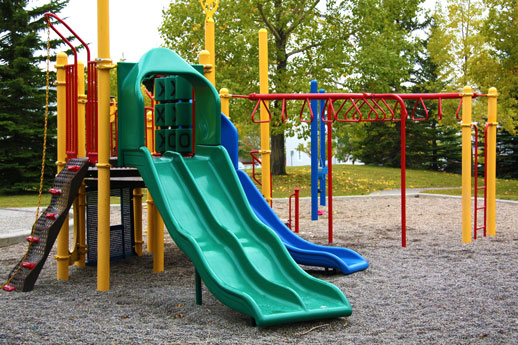 A colorful outdoor children's playground with a green wavy slide, a blue slide, and red and yellow climbing structures on a gravel surface, surrounded by trees.