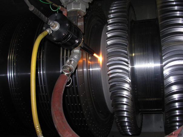 Close-up of a welding torch sparking beside red and yellow hoses and corrugated metal ducts on industrial equipment.