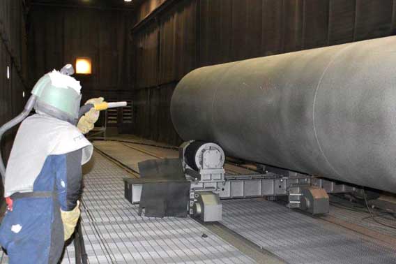 A worker in protective gear stands beside a large cylindrical metal pipe on a roller conveyor inside an industrial facility.