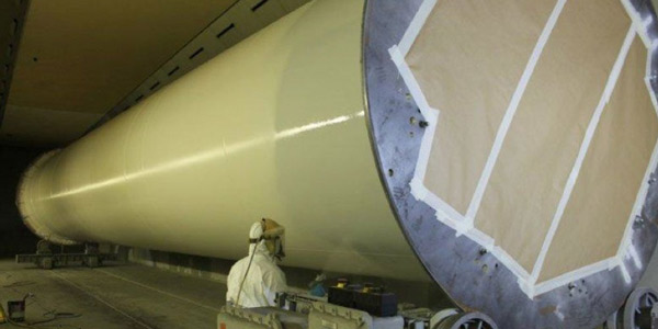 A large rocket fuselage section in a factory hangar with a worker in protective clothing nearby.
