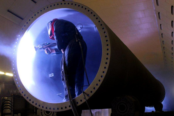 Technician in a protective suit using a tool inside the circular opening of a large rocket component, with blue illumination inside.