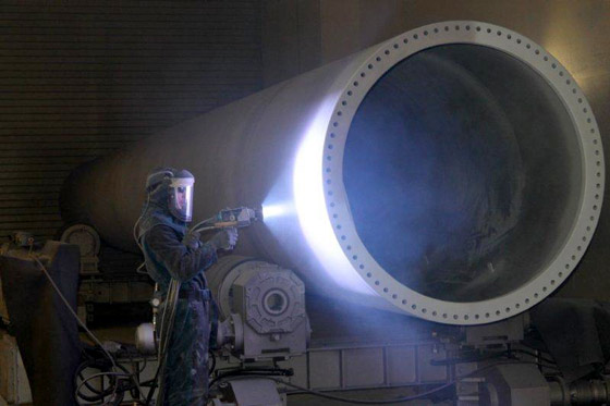 A worker in protective gear and a helmet stands beside a large hollow cylindrical metal tube, welding equipment glowing in an industrial workshop.
