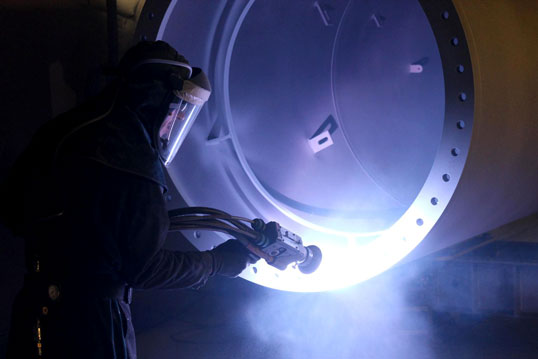 Industrial worker in protective gear inspecting a large circular metal vessel with a bright blue light.