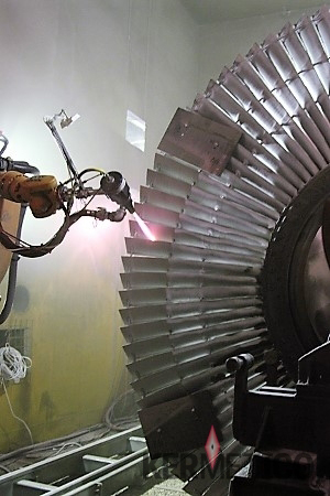 Robotic arm welding near a large jet engine turbine rotor inside an industrial repair facility.