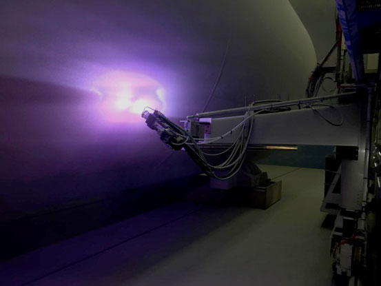 A robotic arm with cables extends toward a purple plasma arc inside a large curved laboratory chamber.