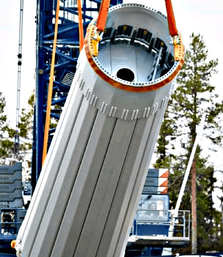 A large gray corrugated cylindrical metal component suspended by orange lifting straps at a construction site with blue scaffolding and trees in the background.
