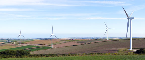 Several wind turbines stand in a patchwork farmland landscape beneath a blue sky.