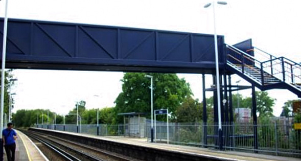 Blue elevated pedestrian footbridge with a right-side stairway crossing over a street, with fencing and trees in the background.