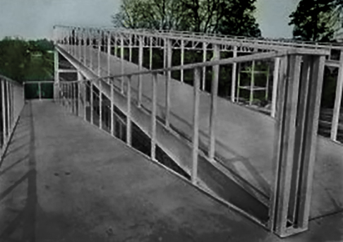 Outdoor construction site with a long metal-framed ramp and guardrails on a concrete surface, trees in the background.