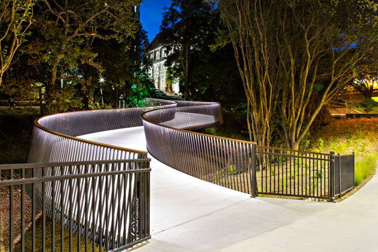 A curved pedestrian bridge with metal railings winds through a park at night toward a lit building.