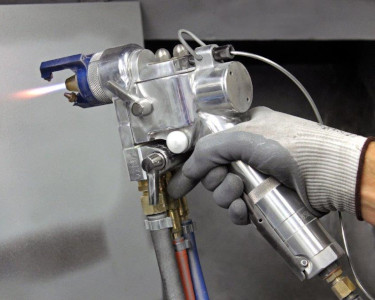 A gloved hand holds a metal spray gun connected to hoses, with a small flame at the nozzle.