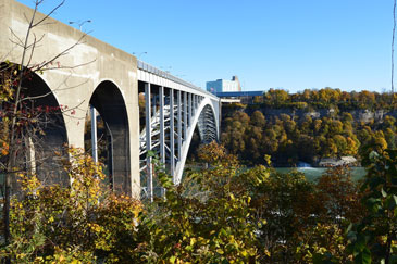 A large arched bridge spans a river beneath a clear blue sky, framed by autumn trees.