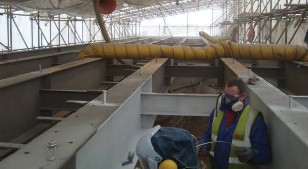 Two workers in PPE inspect large metal panels and yellow ductwork inside an industrial facility.
