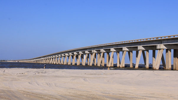 A long concrete pier with evenly spaced supports extends along a sandy beach beneath a clear blue sky.