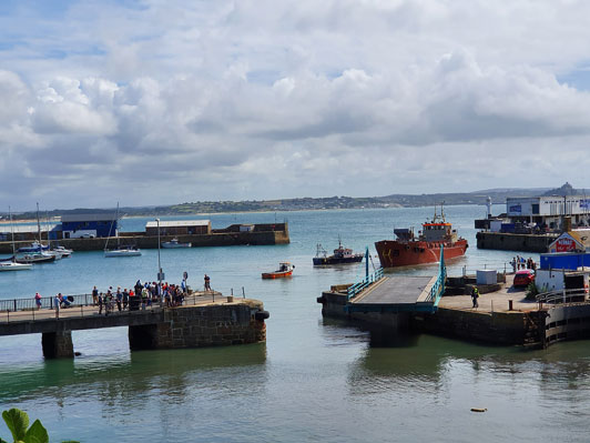 A harbor with several boats docked at concrete piers and people standing along the waterfront under a cloudy sky.