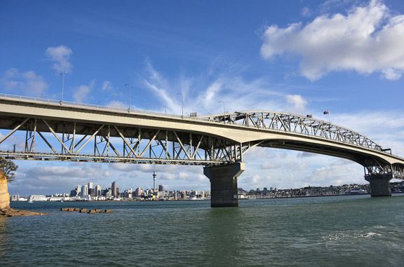 A large steel arch bridge spans a body of water with a distant city skyline beneath a blue sky with clouds.