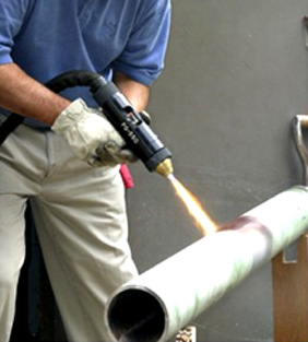 A worker wearing gloves and a blue shirt uses a handheld blowtorch on a large metal pipe.